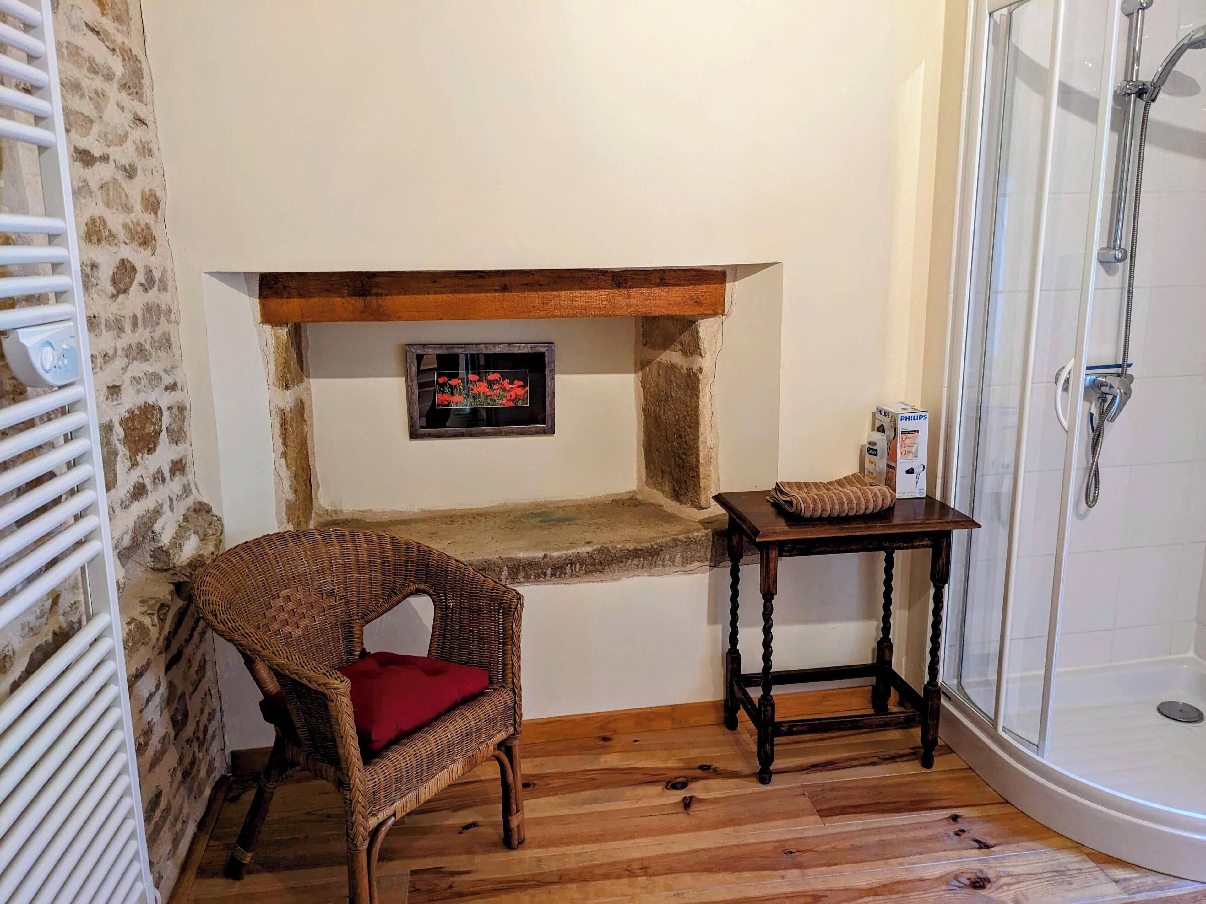 Une petite salle de bain située dans un coin, avec une chaise en bois, une petite table sur laquelle se trouve une boîte, une douche et un mur en pierre.