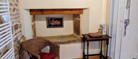 Une petite salle de bain située dans un coin, avec une chaise en bois, une petite table sur laquelle se trouve une boîte, une douche et un mur en pierre.
