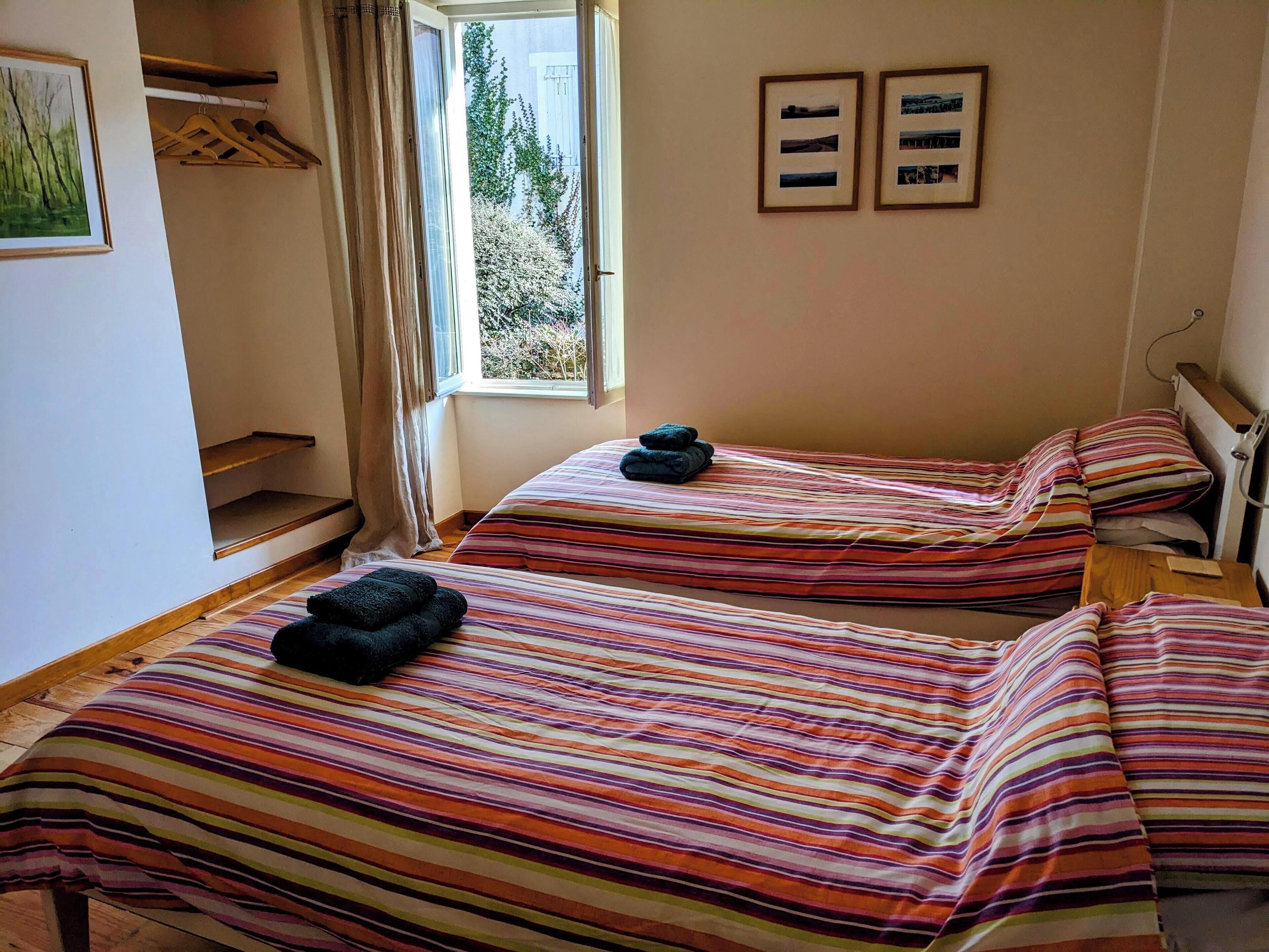 A room with two beds, each with striped bedding and a pair of folded towels. There is a window with curtains, a framed picture on the wall, and a small table with a lamp.