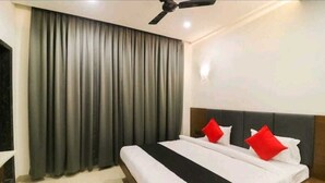 Habitación de hotel con una cama grande, cortinas grises, un ventilador de techo y una mesita de noche con lámpara.