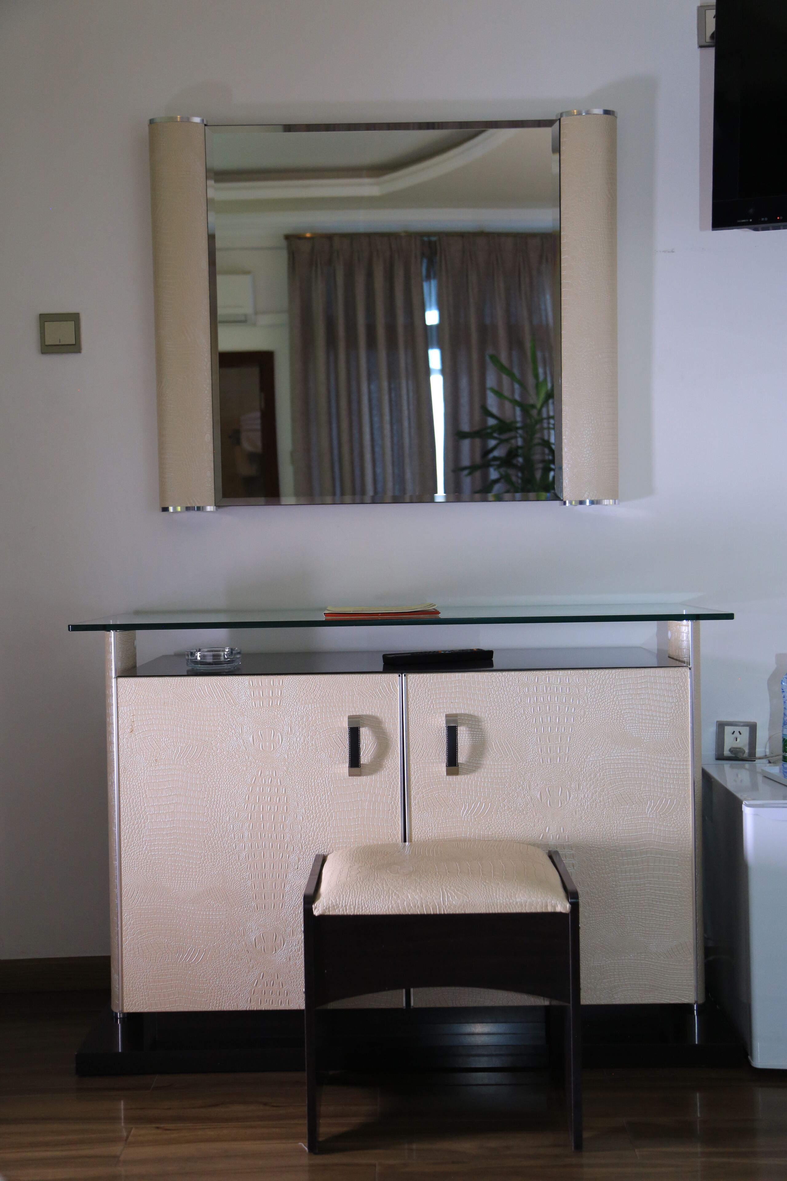 A compact room with a glass-top desk, a beige upholstered chair, and a large mirror above the desk.