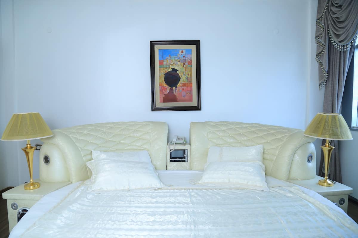 A double bed with a quilted headboard, bedside lamps, a framed artwork on the wall, and a window with curtains.