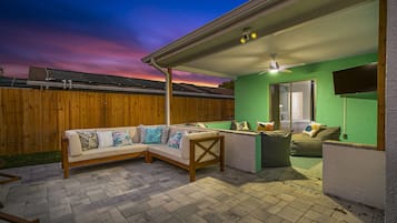 A covered patio area with a wooden fence, a sectional sofa with patterned cushions, and a ceiling fan.