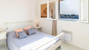 Une chambre à coucher avec un lit, une table de chevet avec une lampe, une fenêtre donnant sur des immeubles et un radiateur.