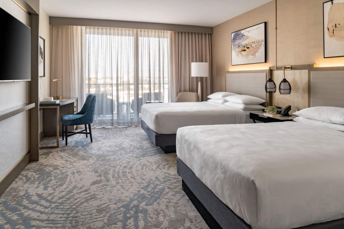 A hotel room with two beds, a desk, a chair, a TV, and a large window with curtains.