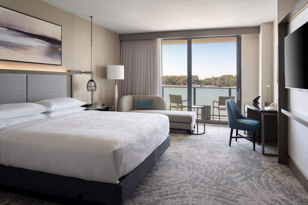 A hotel room with a large bed, a desk, a chair, a TV, and a view of a body of water.