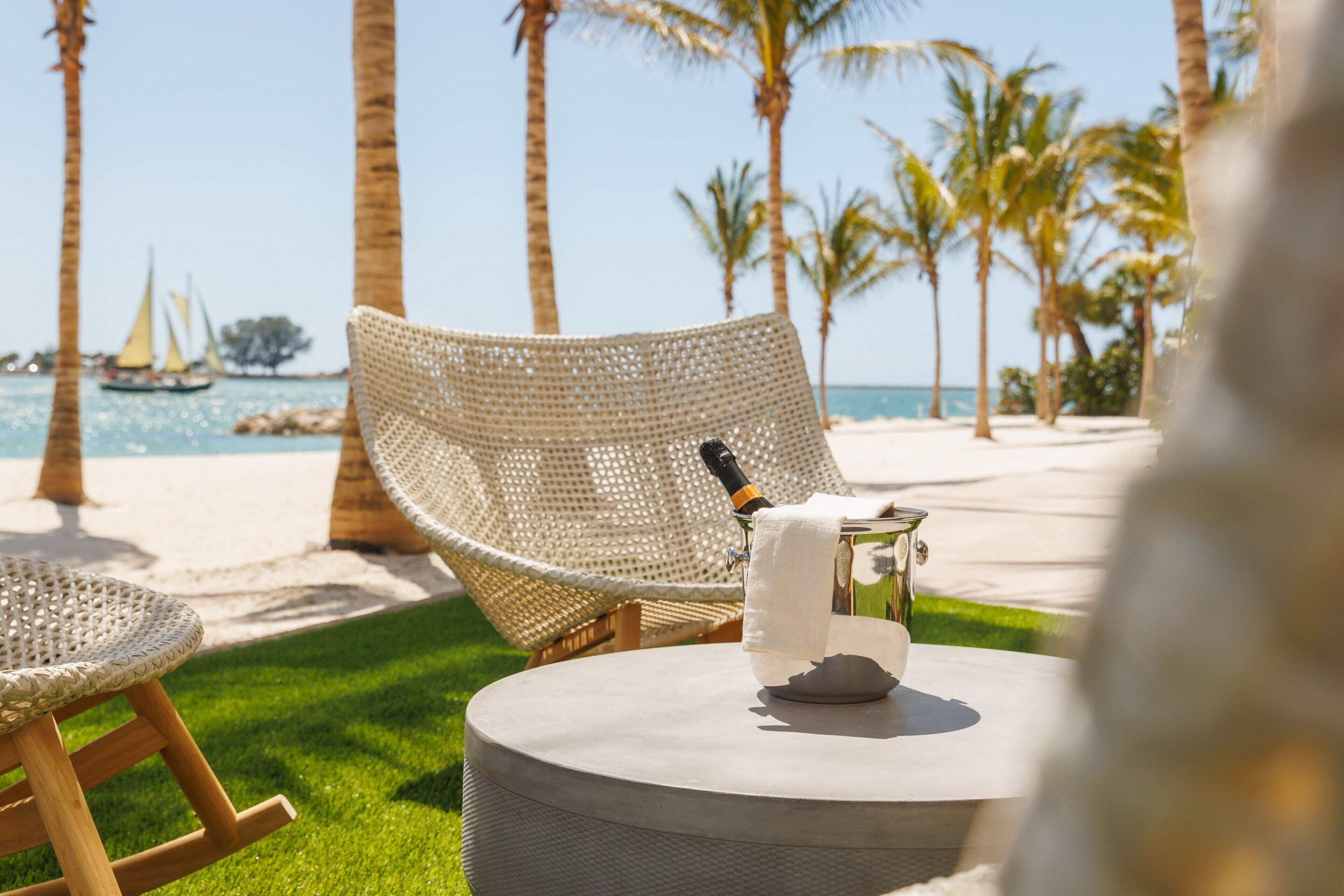 Una silla de mimbre en una mesa redonda, con una botella y un vaso, con vista a una playa con palmeras y un velero a lo lejos.