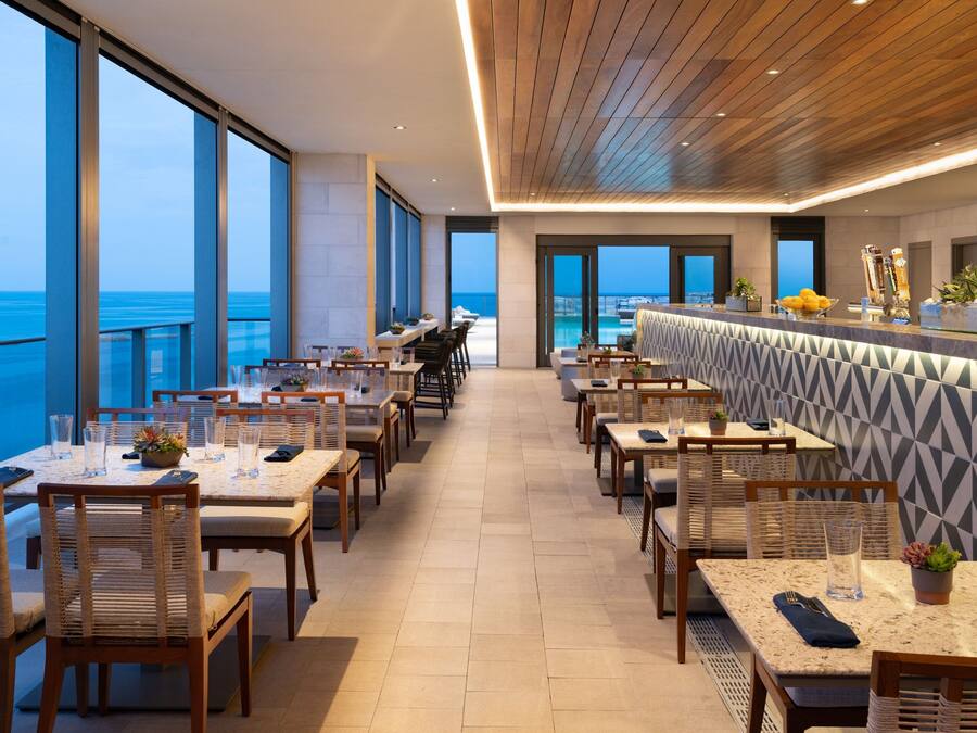 A modern restaurant with a view of the ocean, featuring wooden tables and chairs, a bar area, and large windows.