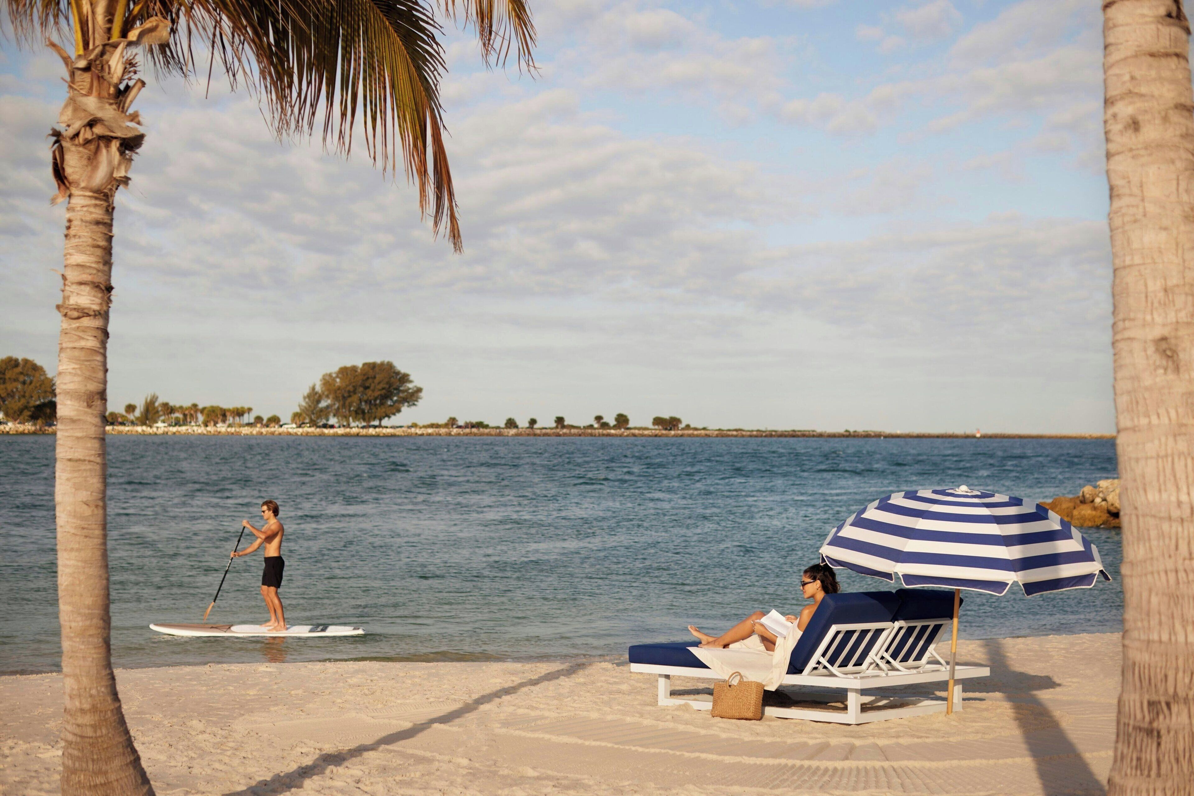 Escena de playa con una persona practicando paddleboard y una silla de playa con sombrilla.