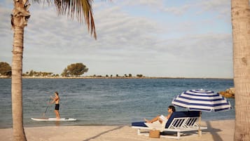 Escena de playa con una persona practicando paddleboard y una silla de playa con sombrilla.