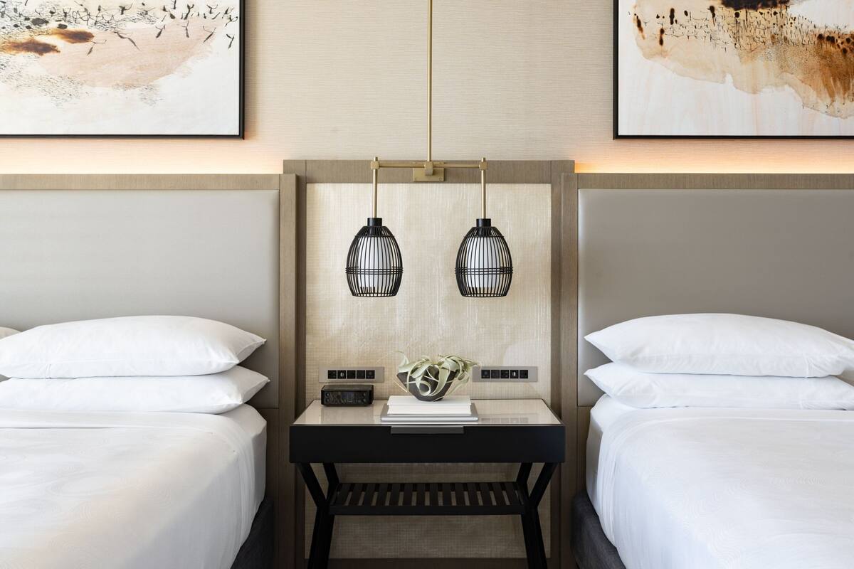 A hotel room with two beds, a nightstand with a lamp, and artwork on the walls.