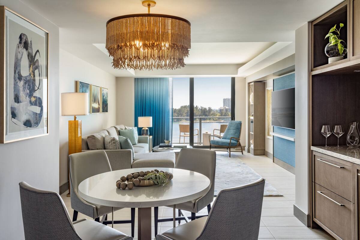 A modern living room with a round dining table, grey upholstered chairs, a chandelier, and a view of the city through large windows.