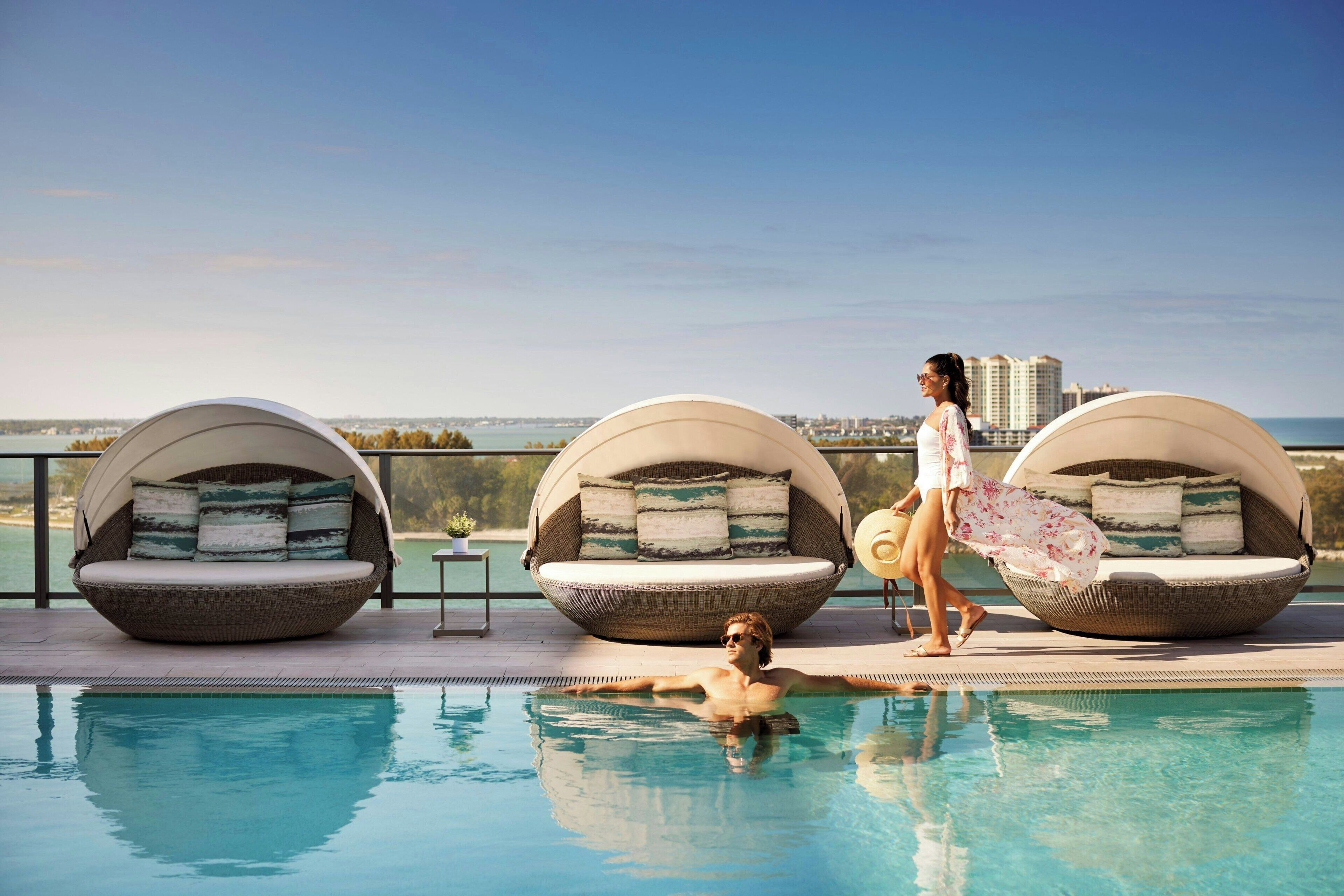 A poolside area with two large, round, wicker lounge chairs with cushions, a clear view of the sea, and a person walking by.