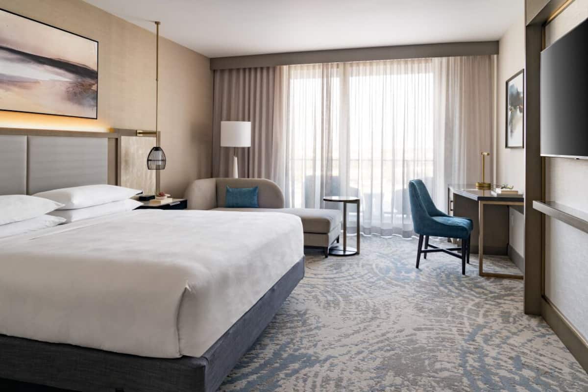 A hotel room with a large bed, a desk, a chair, a lamp, and a TV.