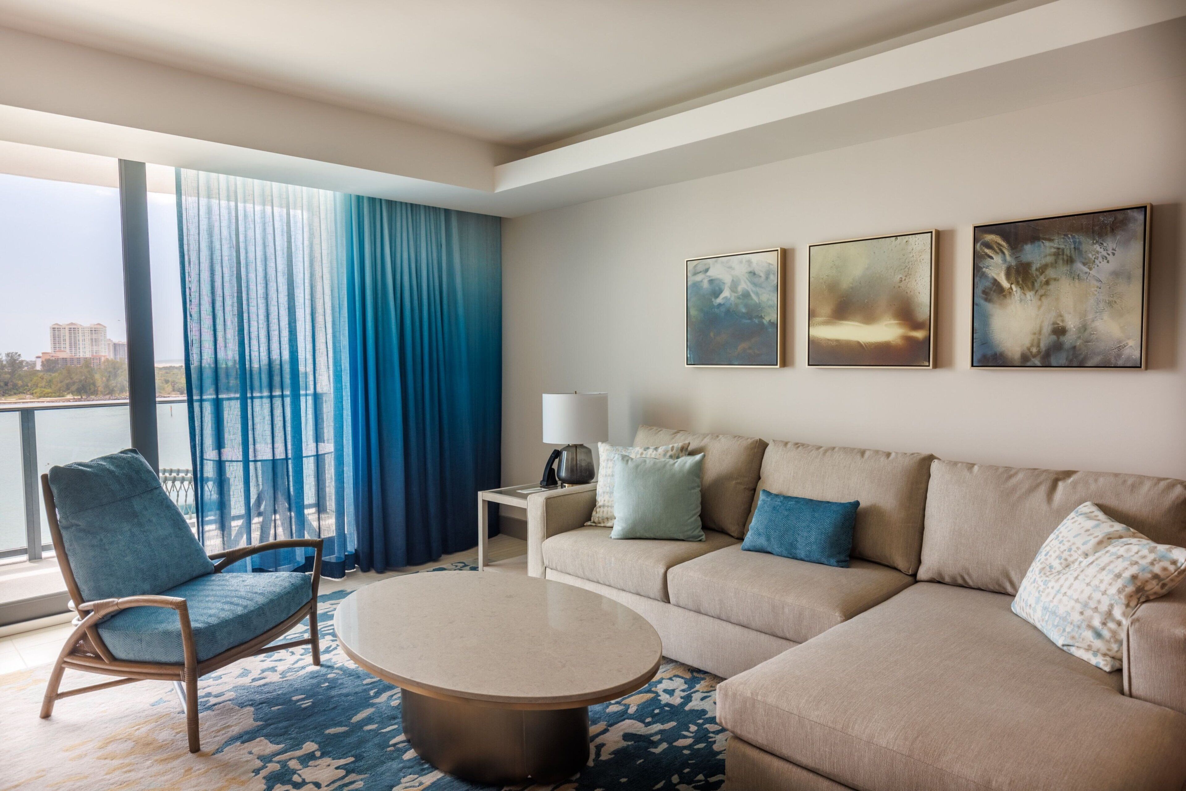Habitación de hotel moderna con un gran ventanal, sofá, sillón, mesa de centro y una obra de arte en la pared.