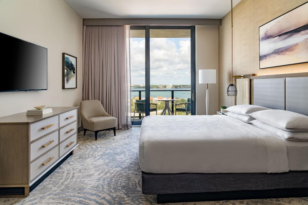 A hotel room with a large bed, a flat-screen TV, a desk with drawers, a chair, and a view of the city through large windows.