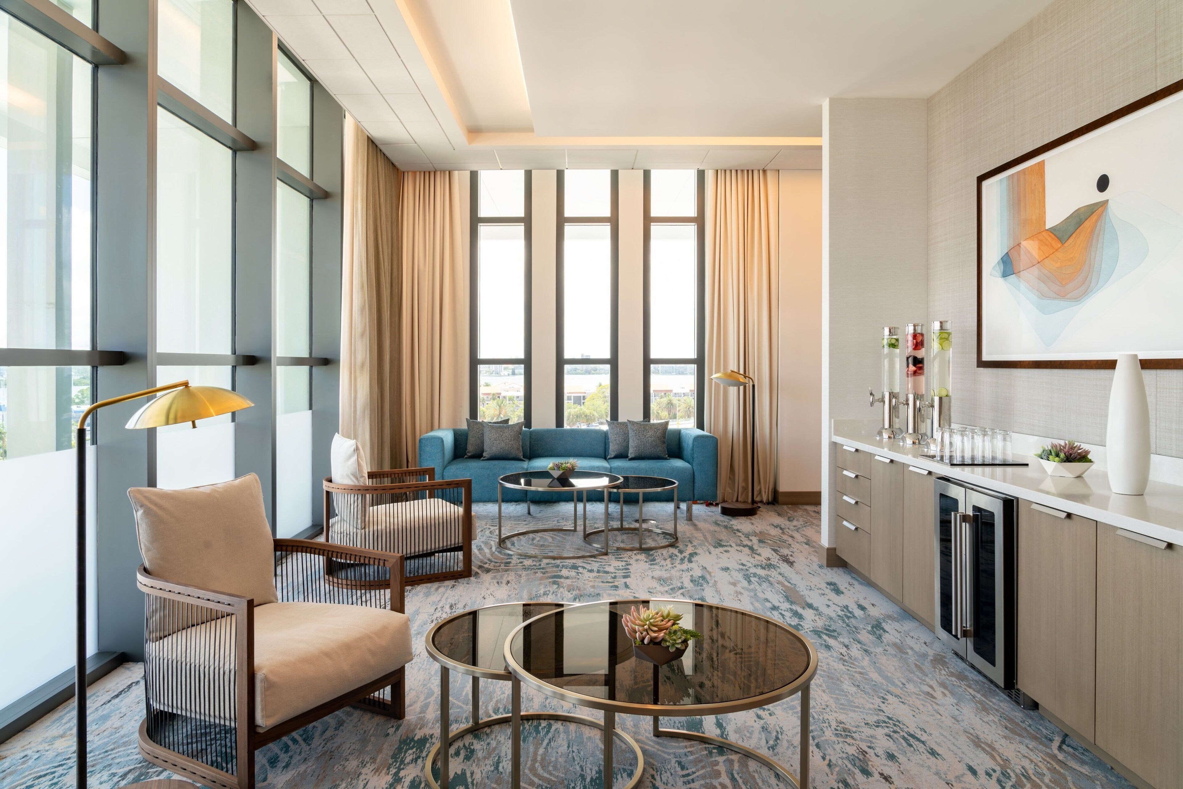 Una habitación de hotel moderna con un sofá azul, una silla de madera, dos mesas redondas, una zona de cocina y ventanas amplias con vistas a la ciudad.