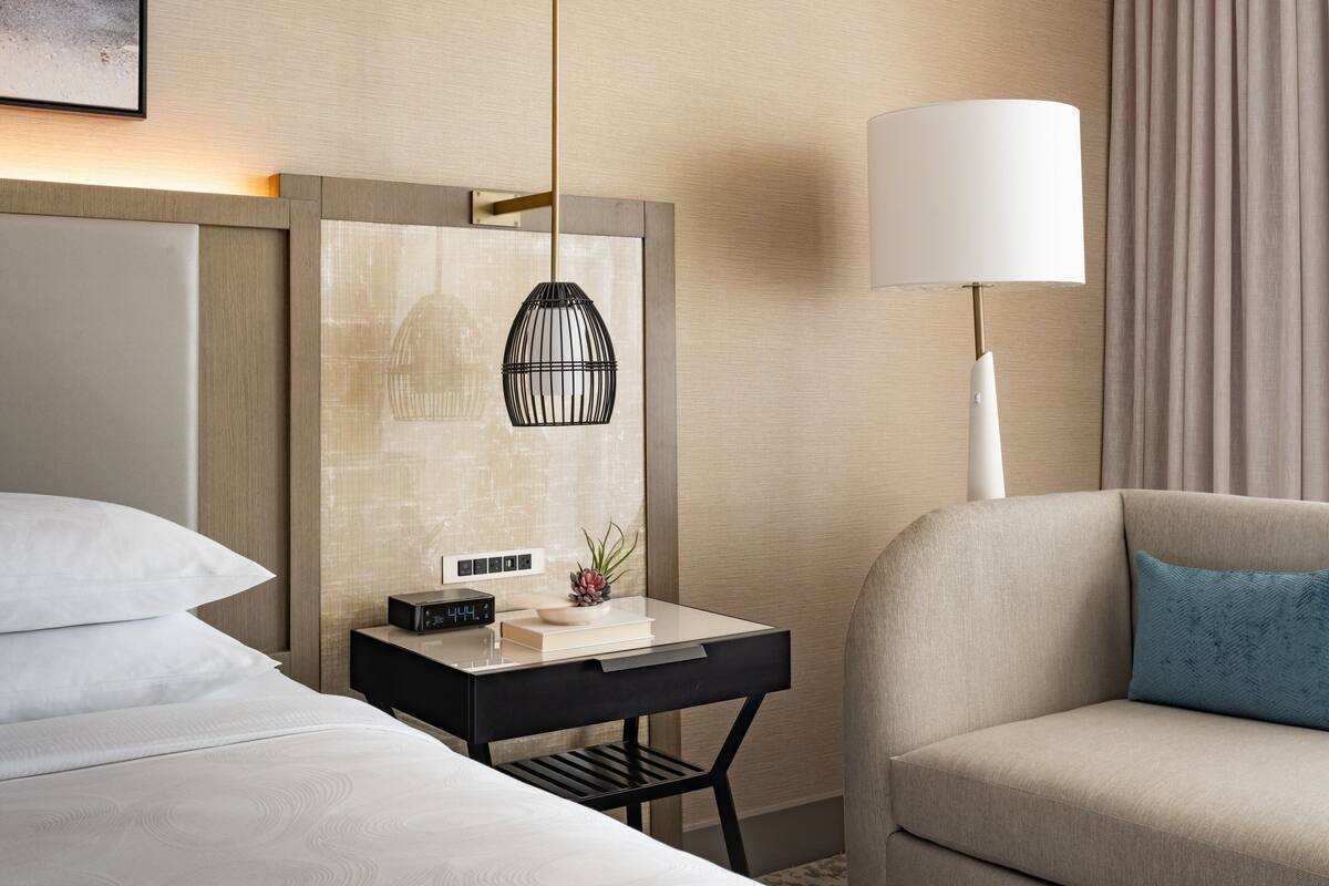 A hotel room with a bed, a desk with a lamp, a sofa, and a wall-mounted artwork.