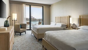 A hotel room with two beds, a desk, a chair, a TV, and a view of the outdoors.