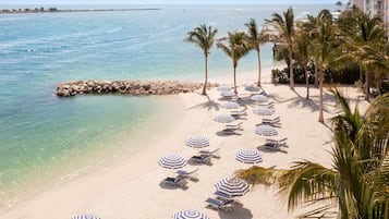 A beach with rows of sun loungers and striped umbrellas, palm trees, and a rocky breakwater.