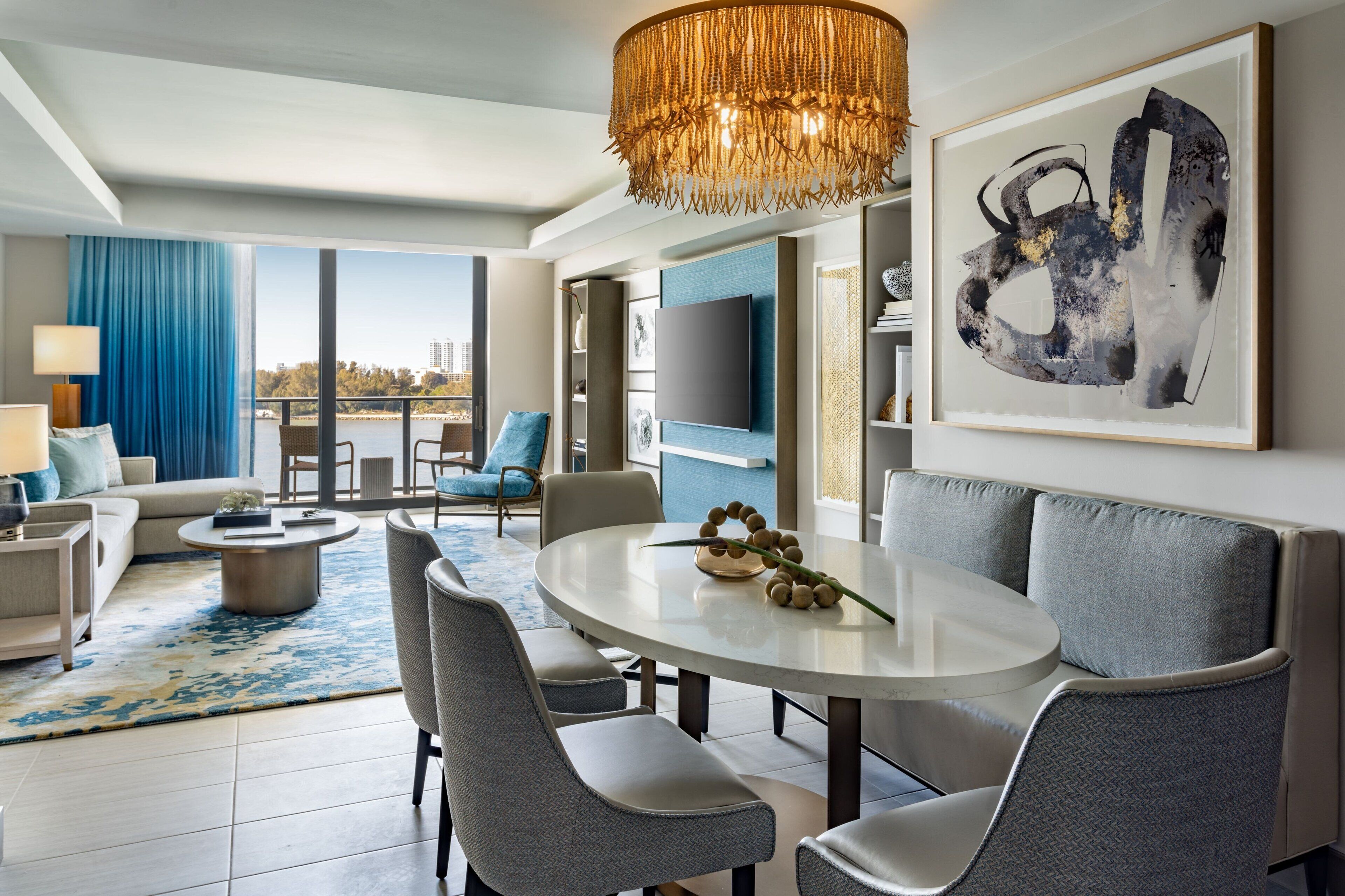 Un salon moderne avec une table à manger ronde, des sièges recouverts d’un tissu gris, un grand tableau abstrait et une vue sur une étendue d’eau.