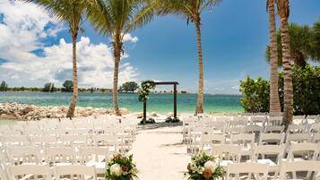 Montaje de boda en la playa con sillas blancas y decoraciones florales, un arco de madera y vista despejada del océano.