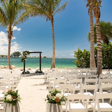 A beach wedding setup with white chairs and floral decorations, a wooden arch, and a clear view of the ocean.