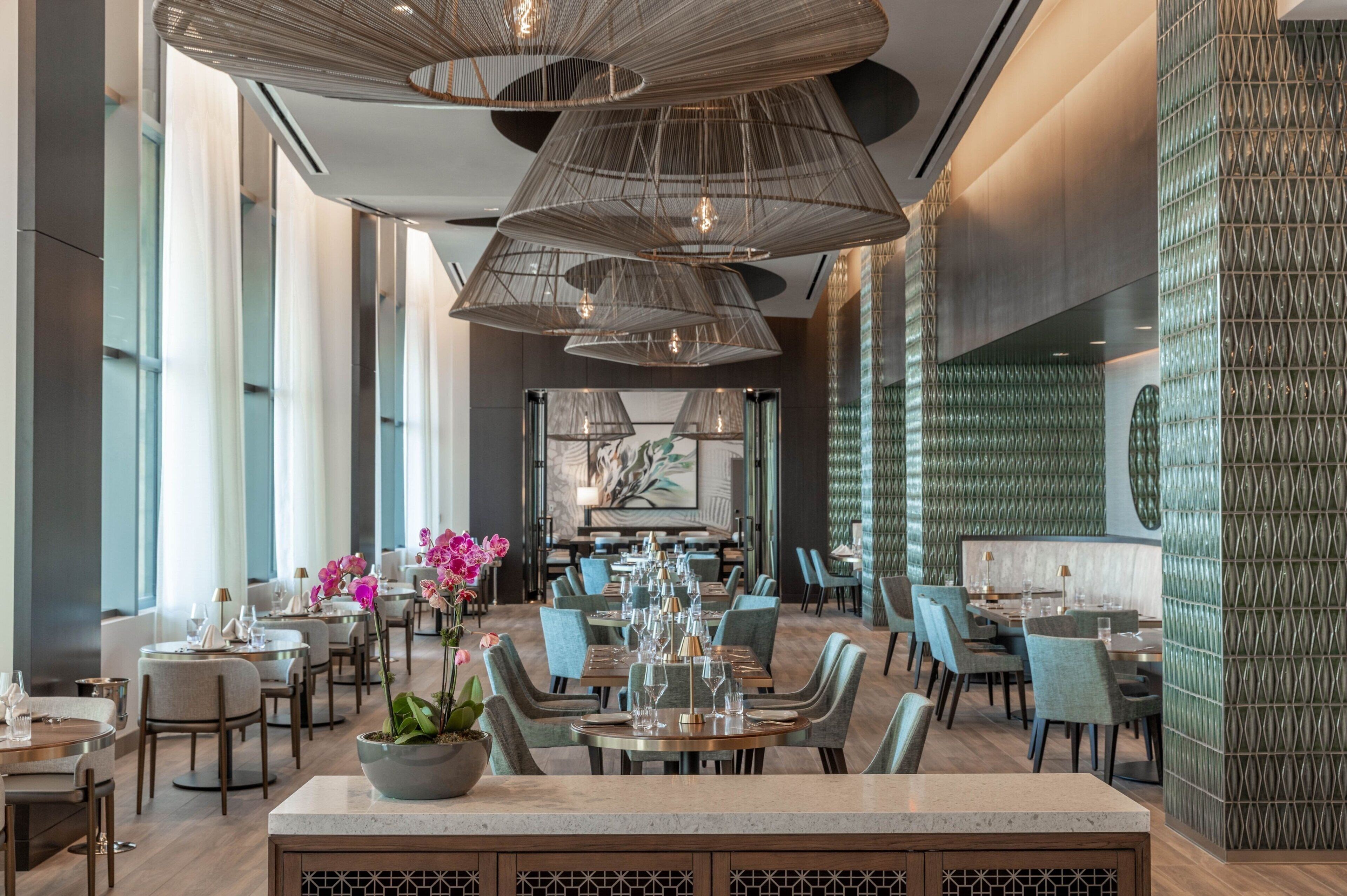 Een modern restaurantinterieur met hoge plafonds, hangende verlichtingsarmaturen en een centraal eetgedeelte met tafels en stoelen.