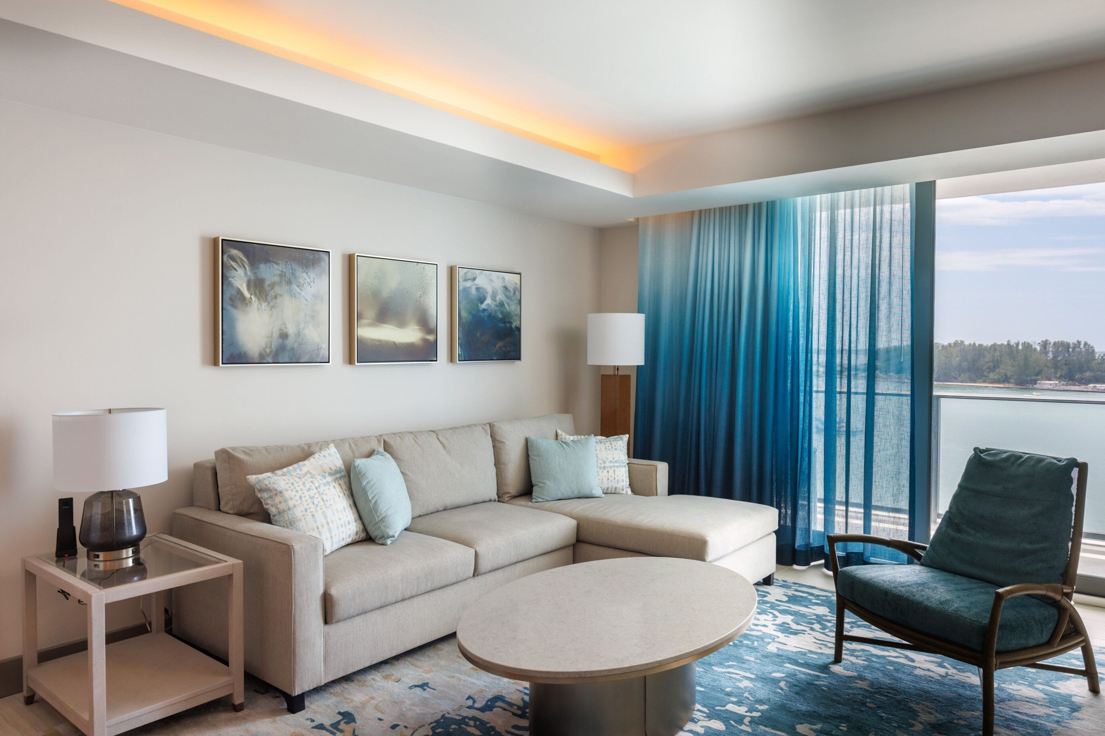 Una habitación de hotel moderna con un sofá seccional color beige, una mesita redonda, una mesita auxiliar con lámpara, y vista al mar a través de ventanas amplias con cortinas azules.