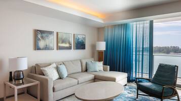 Una habitación de hotel moderna con un sofá seccional color beige, una mesita redonda, una mesita auxiliar con lámpara, y vista al mar a través de ventanas amplias con cortinas azules.