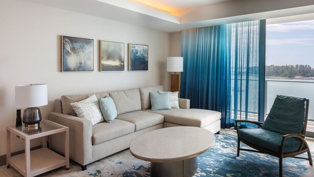 A modern hotel room with a beige sectional sofa, a round coffee table, a side table with a lamp, and a view of the sea through large windows with blue curtains.