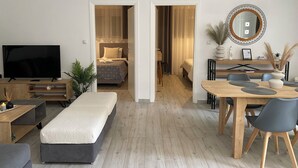 A modern, well-lit bedroom with a large bed, a TV, and a desk area with chairs and a dining table. - Melinta Stay | Apartment near Athens Airport, Rafina Port & Beach (Artemis)