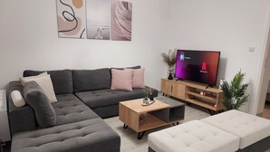 A modern living room with a sectional sofa, a white ottoman, a wooden coffee table, and a flat-screen TV on a wooden stand. - Melinta Stay | Apartment near Athens Airport, Rafina Port & Beach (Artemis)