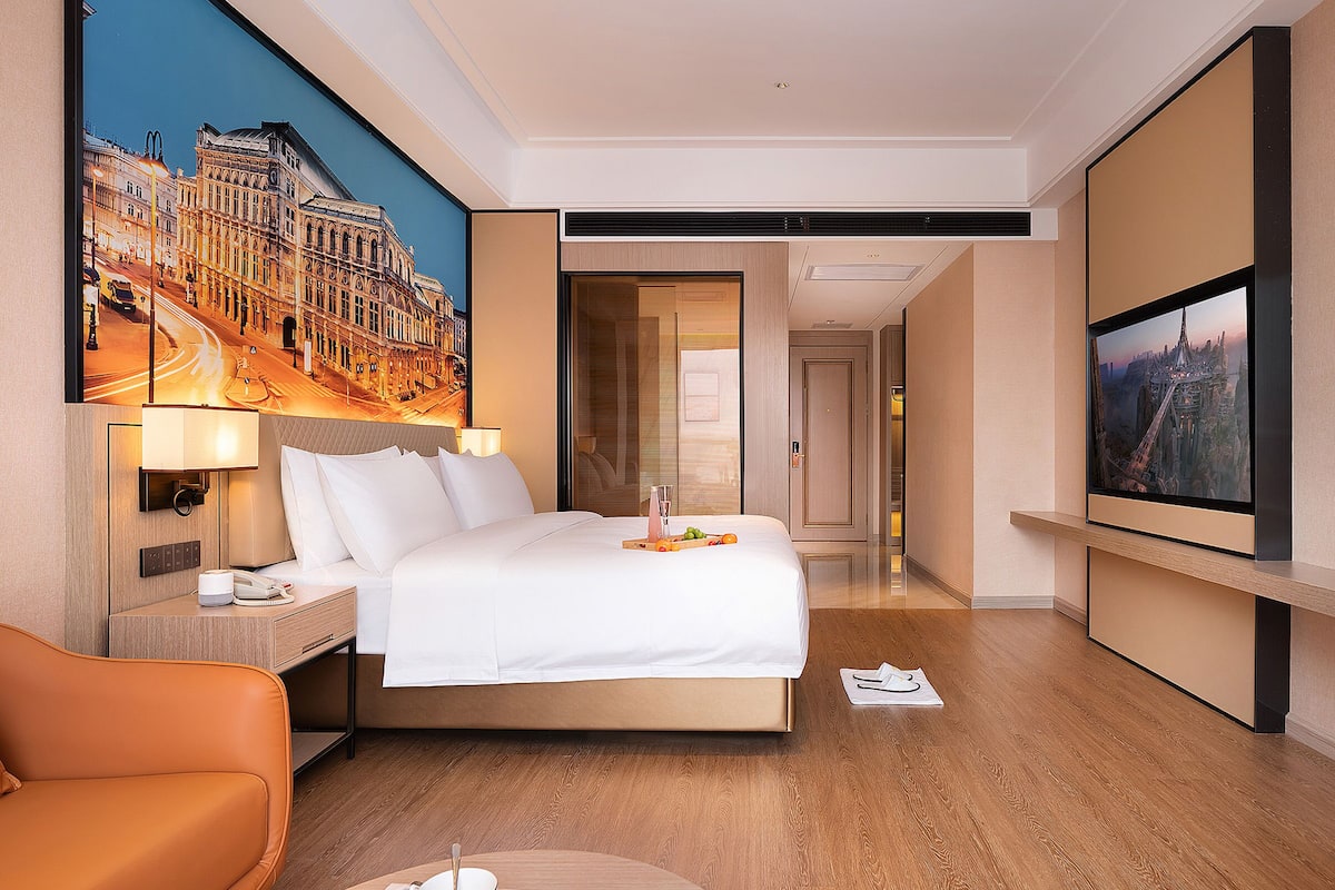 Habitación de hotel con una cama grande, un escritorio con lámpara, una silla, un cuadro de un paisaje urbano y un televisor.