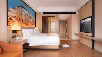 Quarto de hotel com uma cama grande, uma mesa com abajur, uma cadeira, um quadro de uma paisagem urbana e uma televisão.
