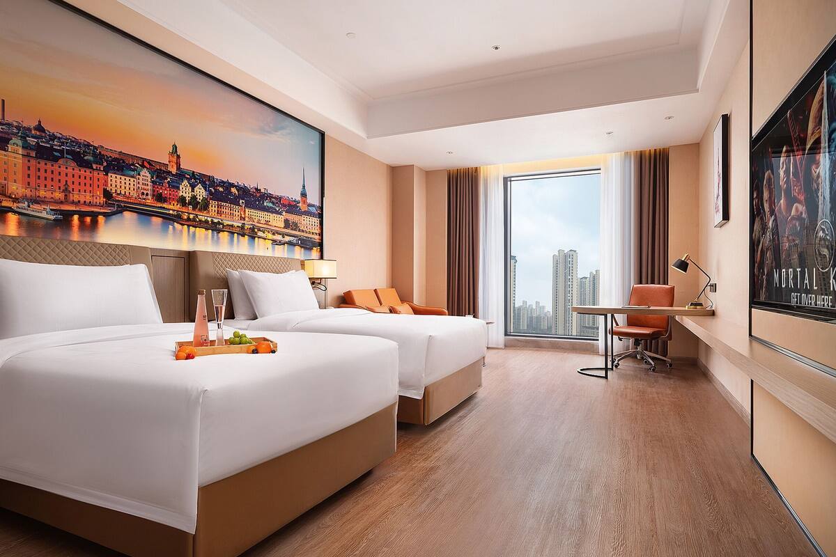 Habitación de hotel con dos camas, un escritorio, una silla, un ventanal con vista a la ciudad y un cuadro en la pared.