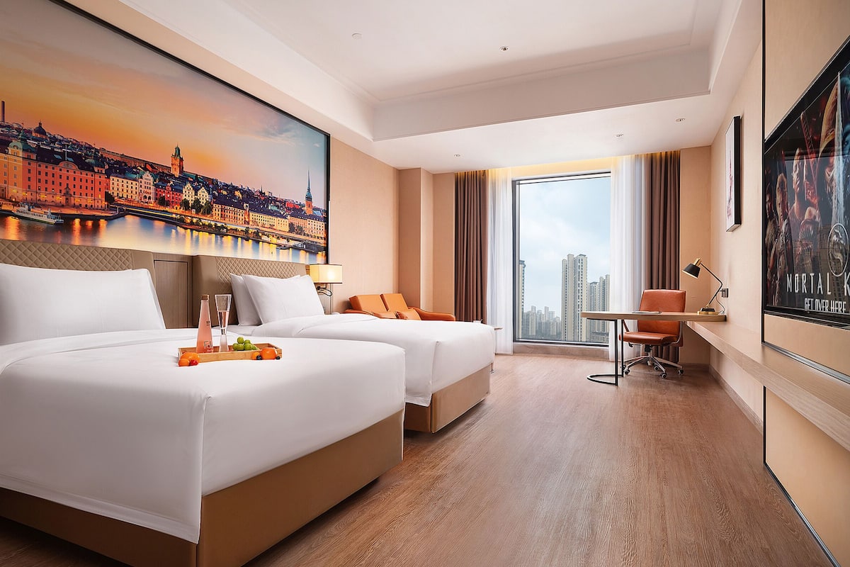Habitación de hotel con dos camas, un escritorio, una silla y un ventanal con vistas a la ciudad.