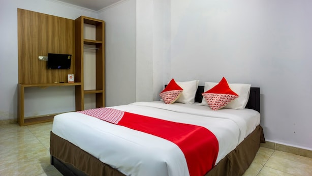 A neatly made bed with a red and white bedspread, two pillows with red and white patterns, and a wooden shelf unit with a television. - Hotel O Wisma Permata Makassar (Makassar)