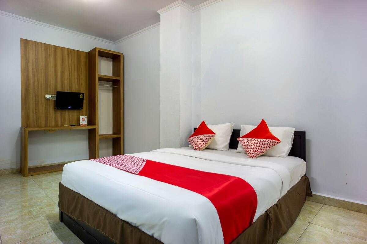 Een netjes opgemaakt bed met een rode en witte spreideken, twee kussens met rode en witte patronen, en een houten plank met een televisie.
