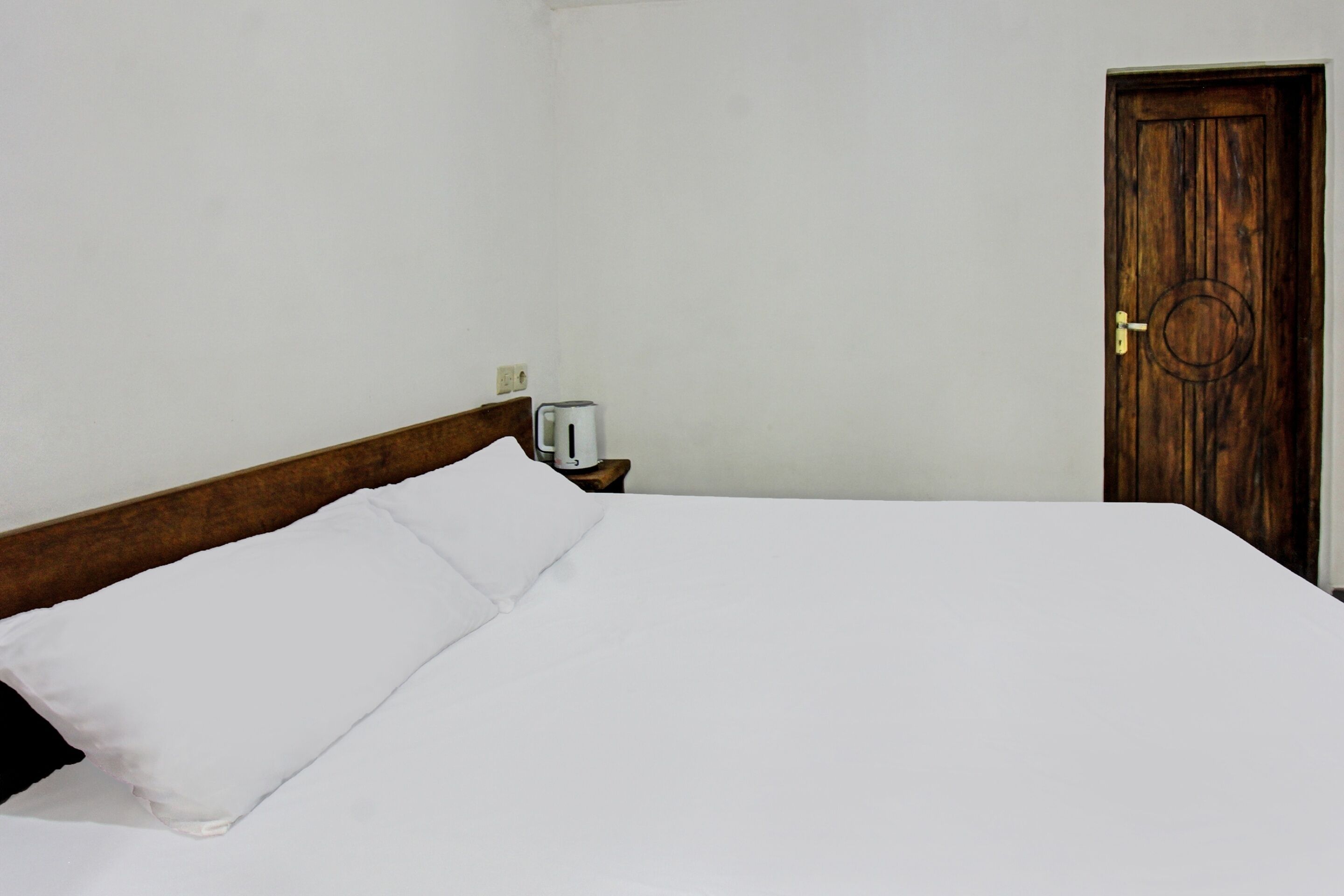 A neatly made bed with white linens in a simple room with a wooden headboard and a wooden door.