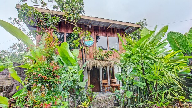 Ein rustikales Holzhaus mit einem strohgedeckten Dach, umgeben von üppiger tropischer Vegetation und einem Bambuszaun.