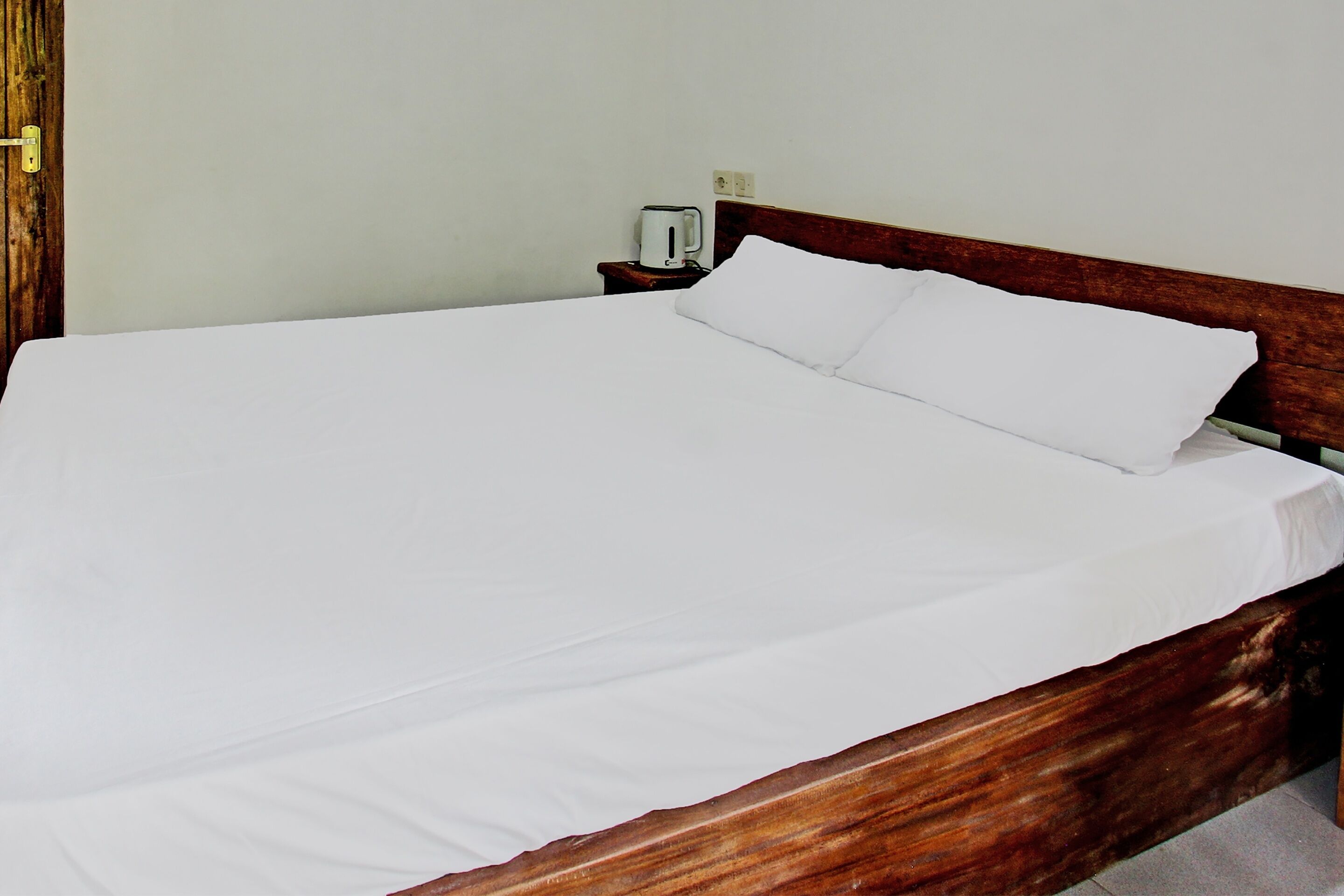 A neatly made bed with white sheets and pillows, a wooden headboard, and a small bedside table with a kettle.
