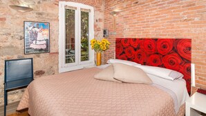 A bedroom with a brick wall, a bed with a red floral headboard, a window with a view of greenery, and a poster on the wall.