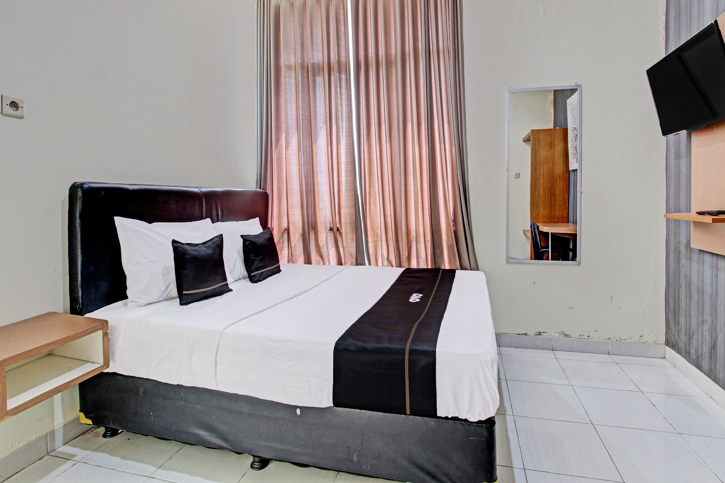 A neatly made bed with a black headboard and white bedding, a wooden nightstand, a mirror, and a TV mounted on the wall.