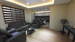 Una sala de estar moderna con un sofá, sillones y una mesa de centro. Se observa una cocina al fondo con una mesa de comedor y sillas.