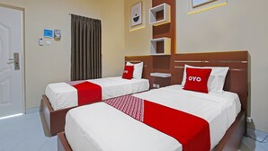 A hotel room with two beds, each with a red and white bedspread and pillows, a wooden headboard, a bedside table, and a wall-mounted shelf with framed pictures. - Capital O 92105 A'shanti Inn (Cilacap)