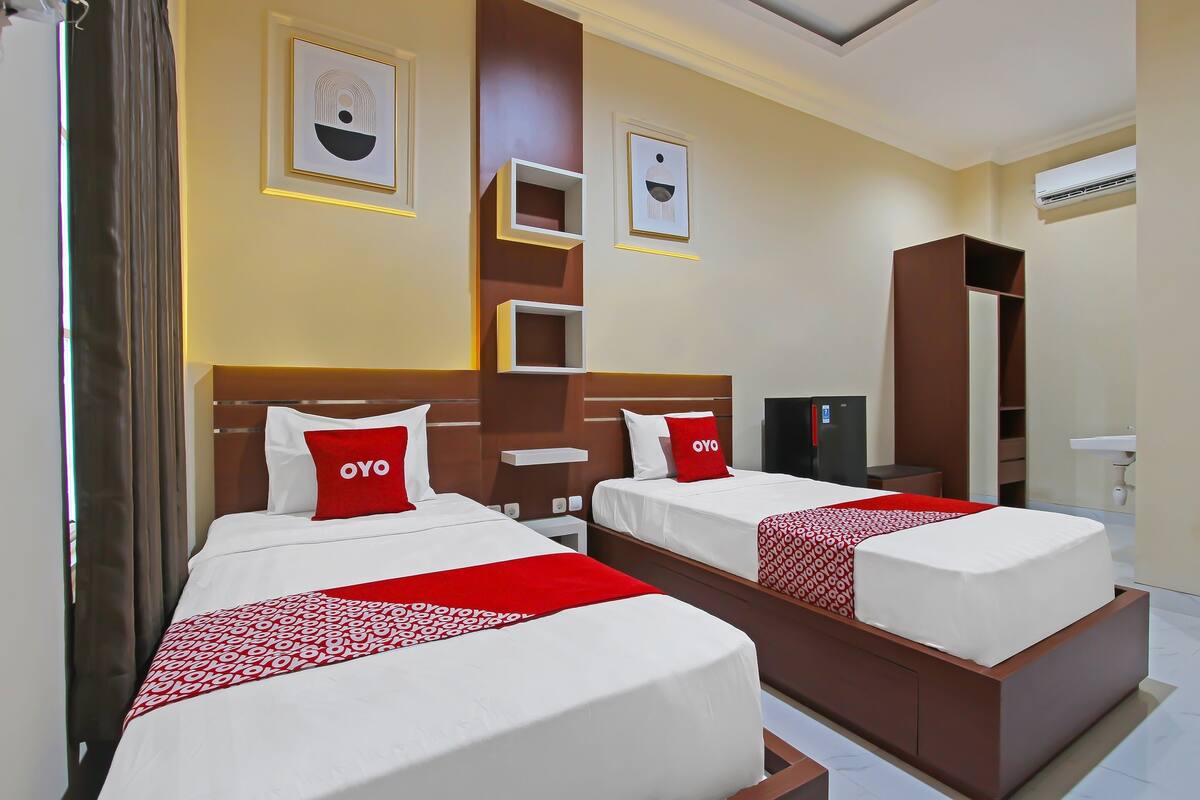 Quarto de hotel com duas camas, cada uma com colcha vermelha e branca, cabeceira de madeira e travesseiro vermelho com o nome da marca 'OYO'. Há prateleiras na parede e um guarda-roupa.