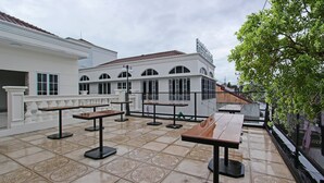 An outdoor patio with tables and benches, a white building with arched windows, and a tree. - Capital O 92105 A'shanti Inn (Cilacap)