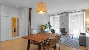 A modern living room with a wooden dining table, wicker chairs, a grey sofa, and a large window with sheer curtains.
