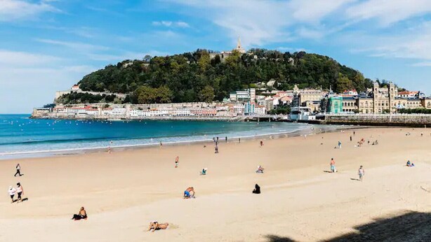 Ein Strand mit badenden und sonnenden Menschen, ein Hügel mit Gebäuden und ein klarer Himmel.